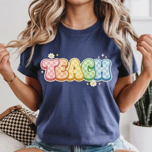May include: Navy blue t-shirt with the word "TEACH" in a colorful, retro font. The letters are outlined in white and have a checkered pattern in shades of pink, orange, yellow, green, and blue. Small white daisies and stars accent the design.