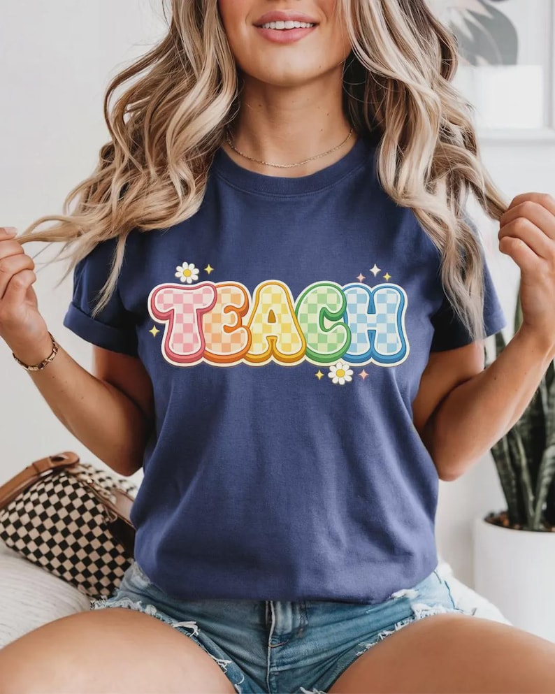 May include: Navy blue t-shirt with the word "TEACH" in a colorful, retro font. The letters are outlined in white and have a checkered pattern in shades of pink, orange, yellow, green, and blue. Small white daisies and stars accent the design.