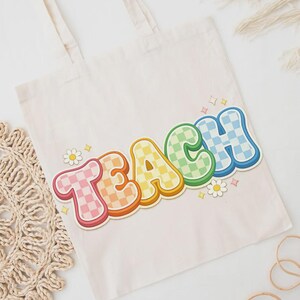 May include: Cream-colored tote bag with the word "TEACH" in colorful, retro-style lettering. The letters are outlined in a darker color and have a checkered pattern in various colors. Small daisy and star graphics are also present.