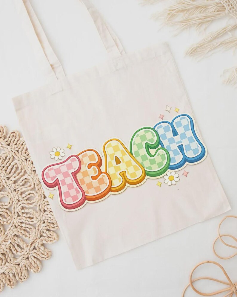 May include: Cream-colored tote bag with the word "TEACH" in colorful, retro-style lettering. The letters are outlined in a darker color and have a checkered pattern in various colors. Small daisy and star graphics are also present.