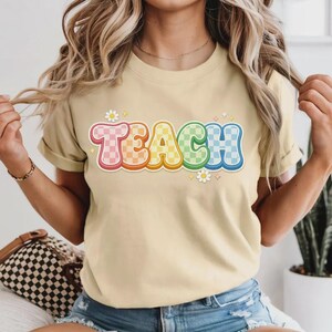 May include: A light tan t-shirt with the word "TEACH" in a colorful, retro font. The letters are outlined in a rainbow gradient and have a checkered pattern. Small daisy graphics are placed around the word.