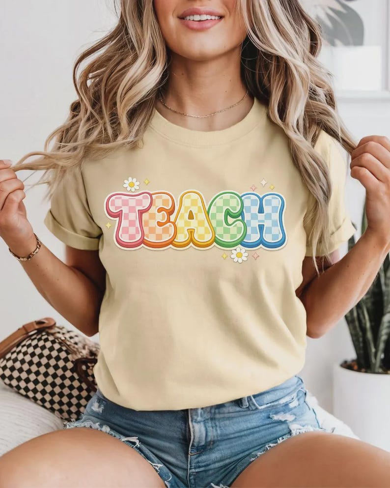 May include: A light tan t-shirt with the word "TEACH" in a colorful, retro font. The letters are outlined in a rainbow gradient and have a checkered pattern. Small daisy graphics are placed around the word.