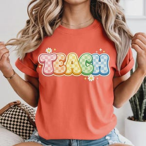 May include: Coral-colored t-shirt with the word "TEACH" in a colorful, checkered design. The letters are outlined in white with a 3D effect, and small daisy graphics are placed around the word. The shirt has rolled-up sleeves.