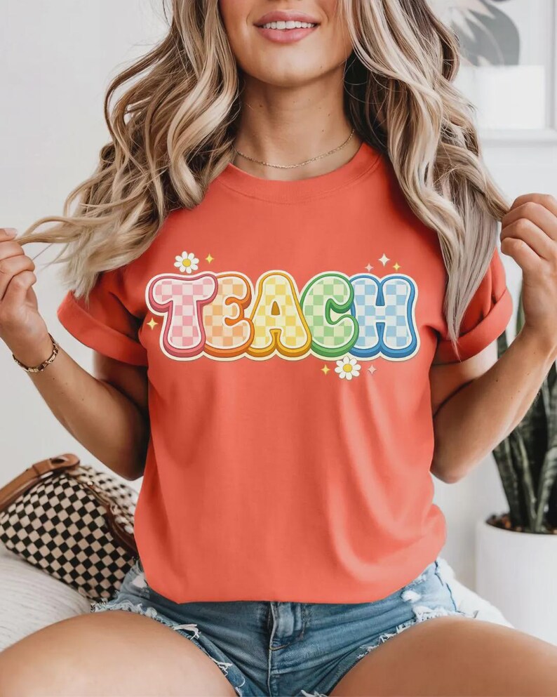 May include: Coral-colored t-shirt with the word "TEACH" in a colorful, checkered design. The letters are outlined in white with a 3D effect, and small daisy graphics are placed around the word. The shirt has rolled-up sleeves.