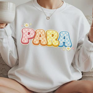Może przedstawiać: Biała bluza z napisem "PARA" w kolorowej, retro czcionce. Litery są r&oacute;żowe, pomarańczowe, ż&oacute;łte i niebieskie, z wzorem w szachownicę. Małe grafiki kwiat&oacute;w i gwiazd na bluzie. Styl casual.