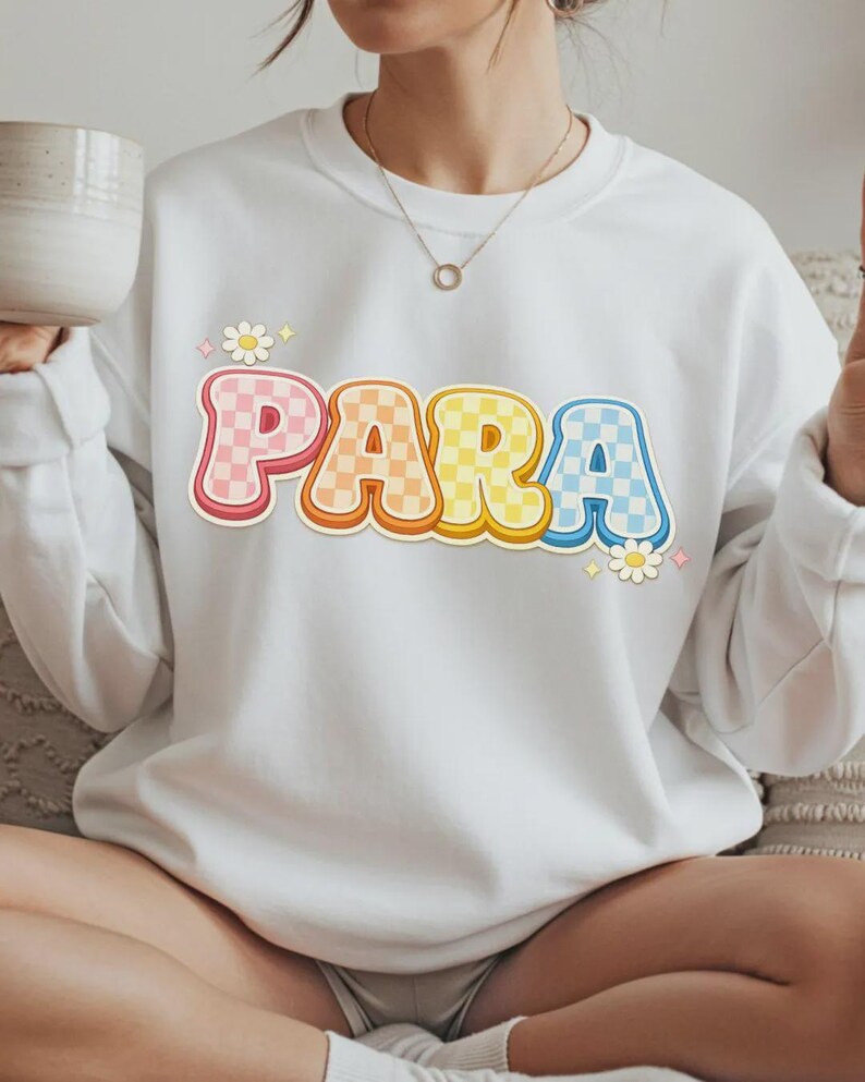 Może przedstawiać: Biała bluza z napisem "PARA" w kolorowej, retro czcionce. Litery są r&oacute;żowe, pomarańczowe, ż&oacute;łte i niebieskie, z wzorem w szachownicę. Małe grafiki kwiat&oacute;w i gwiazd na bluzie. Styl casual.