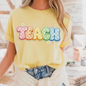 May include: A yellow t-shirt with the word "TEACH" in a colorful, retro font. The letters are outlined in a rainbow of colors and have a checkered pattern. Small daisy and star graphics are placed around the word.