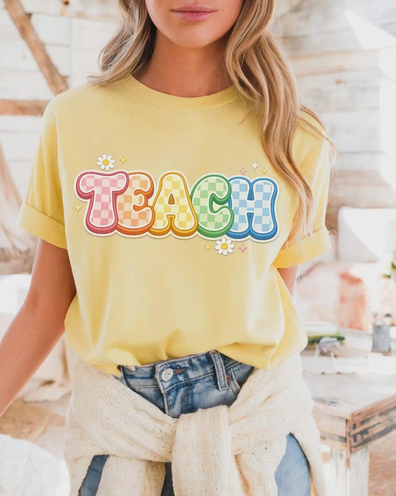 May include: A yellow t-shirt with the word "TEACH" in a colorful, retro font. The letters are outlined in a rainbow of colors and have a checkered pattern. Small daisy and star graphics are placed around the word.