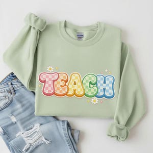 May include: A light green sweatshirt with the word "TEACH" in a colorful, checkered font. The sweatshirt is paired with light blue jeans. The word "TEACH" is in a retro style with a rainbow of colors.