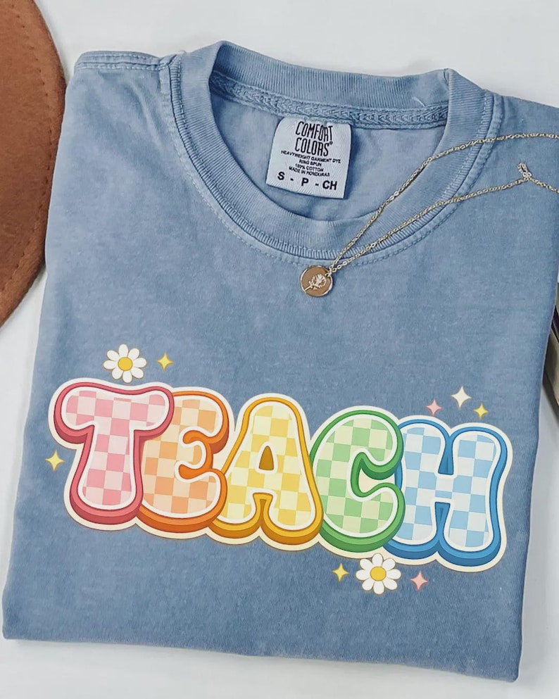 May include: A light blue t-shirt with the word "TEACH" in a colorful, retro font. The letters have a checkered pattern in pink, orange, yellow, green, and blue. A gold necklace with a small pendant rests on the shirt.