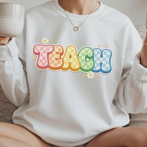 May include: White sweatshirt with the word "TEACH" in a colorful, checkered design. The letters are outlined in a rainbow of colors, with small flower and star accents. A casual, comfortable top for teachers.