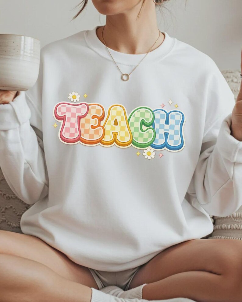 May include: White sweatshirt with the word "TEACH" in a colorful, checkered design. The letters are outlined in a rainbow of colors, with small flower and star accents. A casual, comfortable top for teachers.