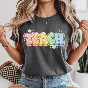 May include: Dark gray t-shirt with the word "TEACH" in a colorful, retro font. The letters are outlined in orange and filled with a checkered pattern in shades of pink, orange, yellow, green, and blue. Small white flowers and stars accent the design.
