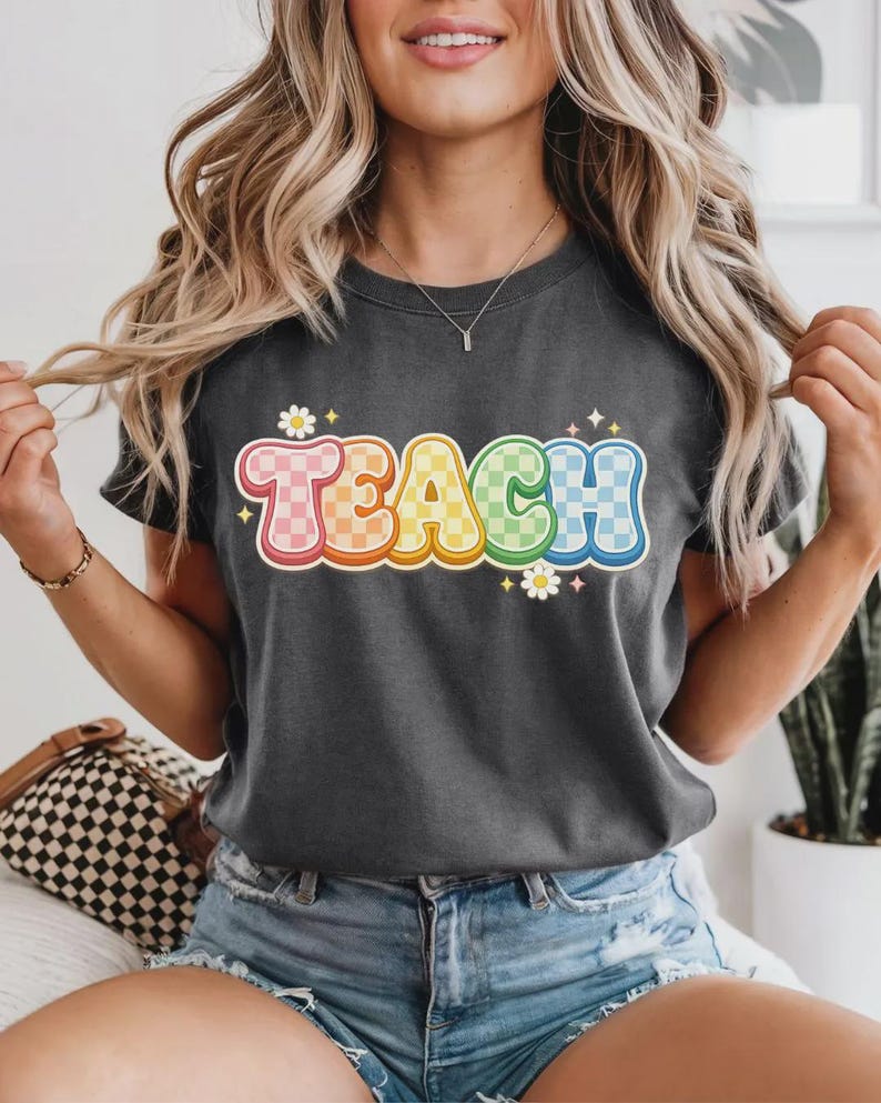 May include: Dark gray t-shirt with the word "TEACH" in a colorful, retro font. The letters are outlined in orange and filled with a checkered pattern in shades of pink, orange, yellow, green, and blue. Small white flowers and stars accent the design.