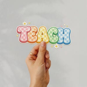 May include: A colorful sticker with the word "TEACH" in a retro font. The letters are in a rainbow of colors with a checkered pattern. Daisies and sparkles surround the word. The sticker is held by a hand.