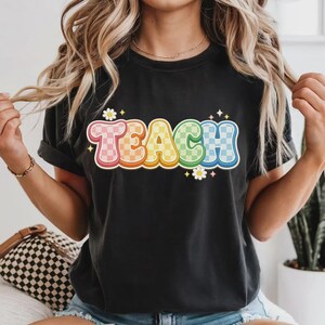 May include: Black t-shirt with the word "TEACH" in colorful, checkered bubble letters. The letters are outlined in white and have a retro, groovy style. Small daisy graphics are placed around the word.
