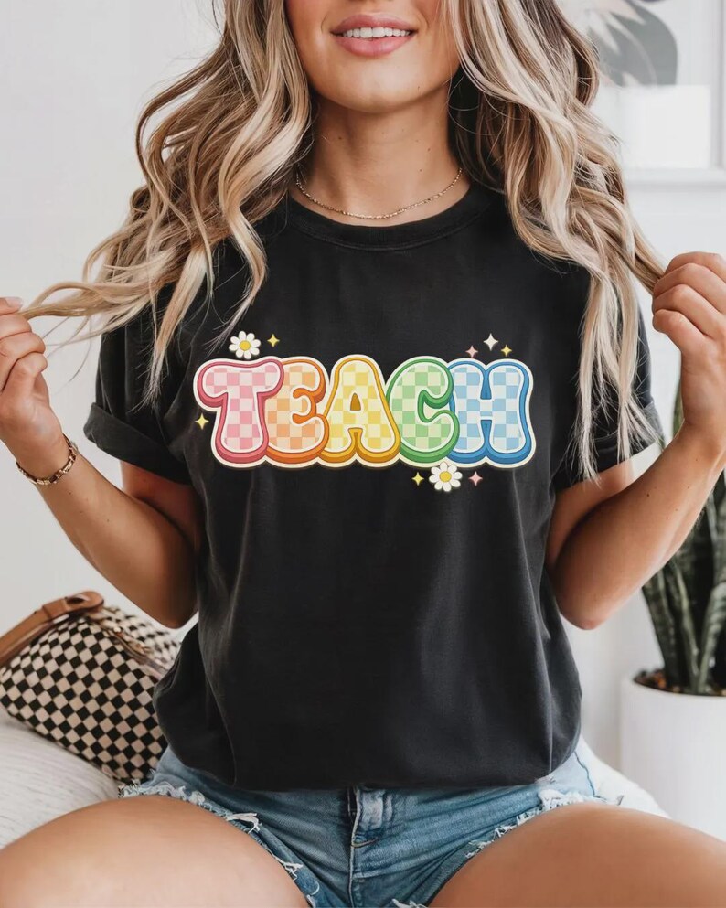 May include: Black t-shirt with the word "TEACH" in colorful, checkered bubble letters. The letters are outlined in white and have a retro, groovy style. Small daisy graphics are placed around the word.