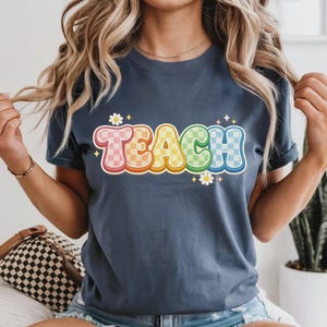 May include: A navy blue t-shirt with the word "TEACH" in a colorful, retro font. The letters are outlined in white and have a checkered pattern in pink, yellow, green, and blue. Small daisy graphics are placed around the text.