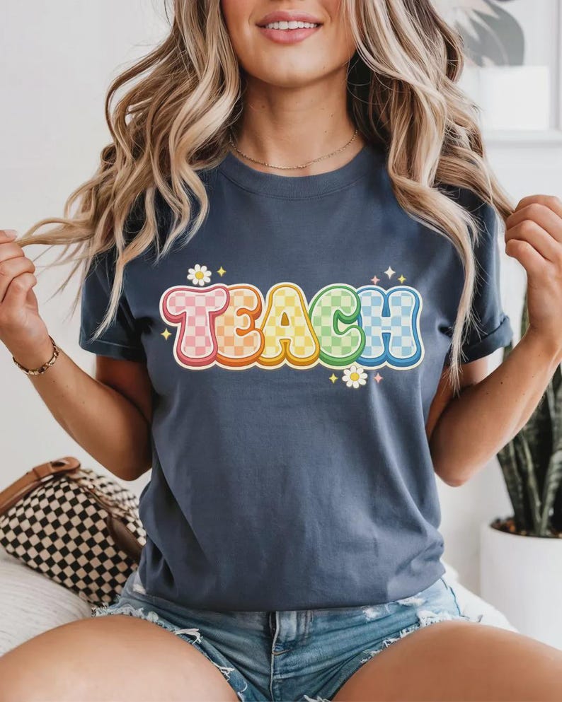 May include: A navy blue t-shirt with the word "TEACH" in a colorful, retro font. The letters are outlined in white and have a checkered pattern in pink, yellow, green, and blue. Small daisy graphics are placed around the text.