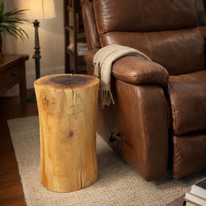 May include: A natural wood stump side table with a dark brown top and light brown sides. The table is cylindrical and sits on a beige rug next to a brown leather recliner.