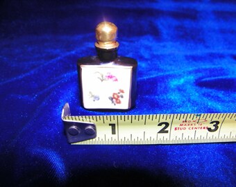 Op de afbeelding: Een klein, vierkant, zwart en gouden parfumflesje met een bloemenontwerp aan de voorkant. Het flesje is ongeveer 2,5 cm hoog en staat op een blauwe ondergrond naast een meetlint.