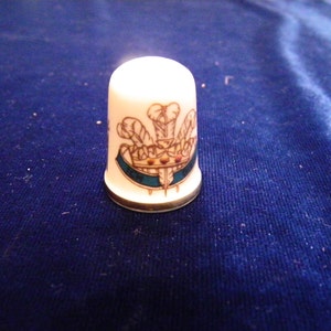 Puede incluir: Dedal de porcelana blanca con ribete dorado y un diseño colorido de una corona y plumas.