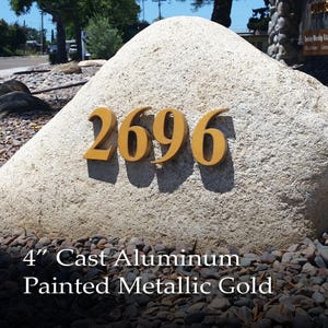 May include: A large, light-colored rock with the number "2696" in gold painted aluminum letters. The text "4" Cast Aluminum Painted Metallic Gold" is below the numbers.