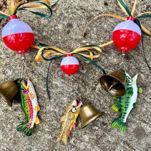May include: Three hanging ornaments featuring red and white spherical fishing bobbers topped with gold and green ribbon bows. Each bobber is attached to a brass-colored metal bell and a small, hand-painted fish figurine in various colors.