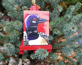 May include: A hand-painted ornament of a loon on a red easel. The loon is black and white with a purple head and red eye. The ornament is signed "AW".