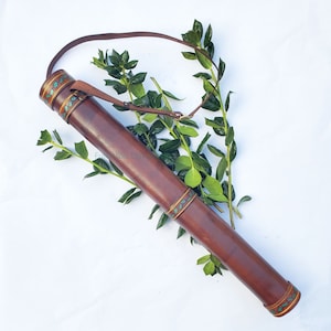 May include: A brown leather quiver with a shoulder strap, decorated with green and gold accents. The cylindrical case is designed to hold arrows. The quiver is shown with green foliage in the background.