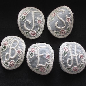 May include: Six embroidered fabric patches with white letters on a light blue background. Each patch has a white letter surrounded by a floral design with pink and green flowers.