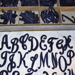 May include: A set of black felt letters in a cursive font. The letters are arranged in alphabetical order, starting with A and ending with P. The letters are on a white surface.