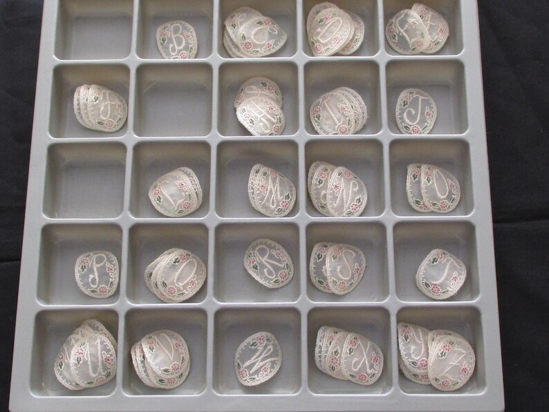 May include: A white plastic tray with 25 compartments, each containing a stack of white lace appliques with embroidered letters. The letters are in a variety of fonts and styles.