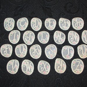May include: A set of 26 embroidered felt letters in a cream color with pink and green floral accents. The letters are arranged in alphabetical order.