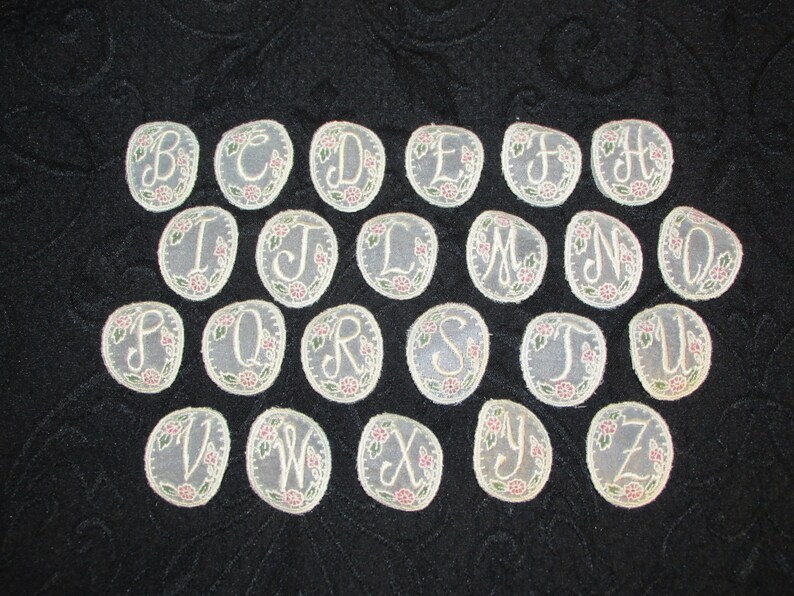 May include: A set of 26 embroidered felt letters in a cream color with pink and green floral accents. The letters are arranged in alphabetical order.