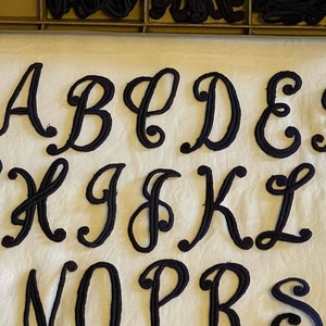 May include: Black felt letters of the alphabet in a cursive font. The letters are arranged in alphabetical order, starting with A and ending with S.
