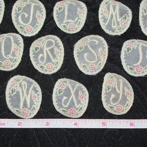 May include: A set of 26 oval-shaped fabric appliques featuring the letters of the alphabet in a white cursive font. Each applique is decorated with a floral design in shades of pink and green.