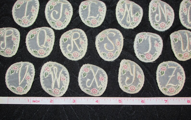 May include: A set of 26 oval-shaped fabric appliques featuring the letters of the alphabet in a white cursive font. Each applique is decorated with a floral design in shades of pink and green.