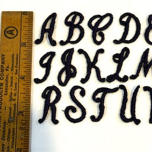 May include: A set of dark navy blue embroidered alphabet letters in a cursive script font. A wooden ruler is positioned vertically on the left side, showing that the letters measure approximately 1.5 inches in height from top to bottom.