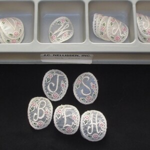 May include: Assortment of oval-shaped, white fabric appliques with embroidered letters and floral designs. The appliques are arranged in a plastic container with individual compartments. The container has a label that reads "J.C. Nellissen, Inc."