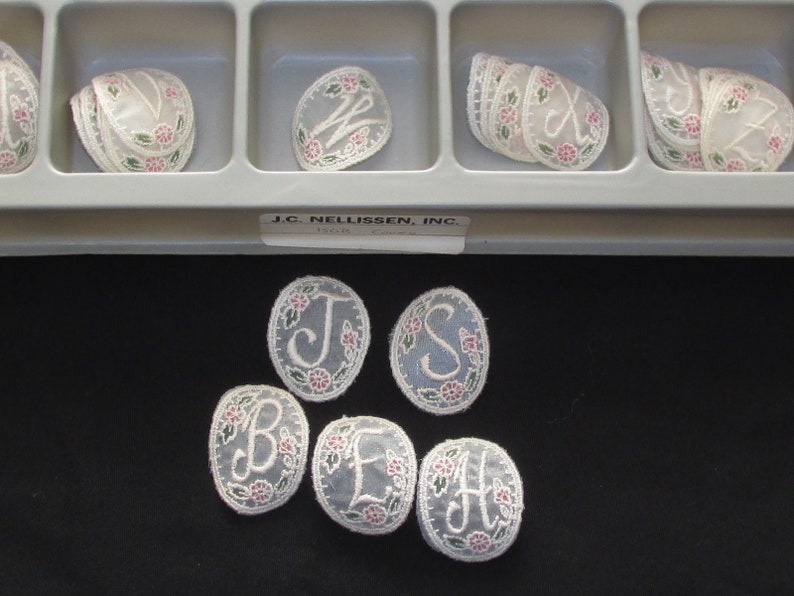 May include: Assortment of oval-shaped, white fabric appliques with embroidered letters and floral designs. The appliques are arranged in a plastic container with individual compartments. The container has a label that reads "J.C. Nellissen, Inc."