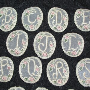 May include: A set of 13 oval-shaped fabric patches with embroidered letters of the alphabet, A through M, in a cream color. The letters are surrounded by a floral design with green leaves and pink flowers.