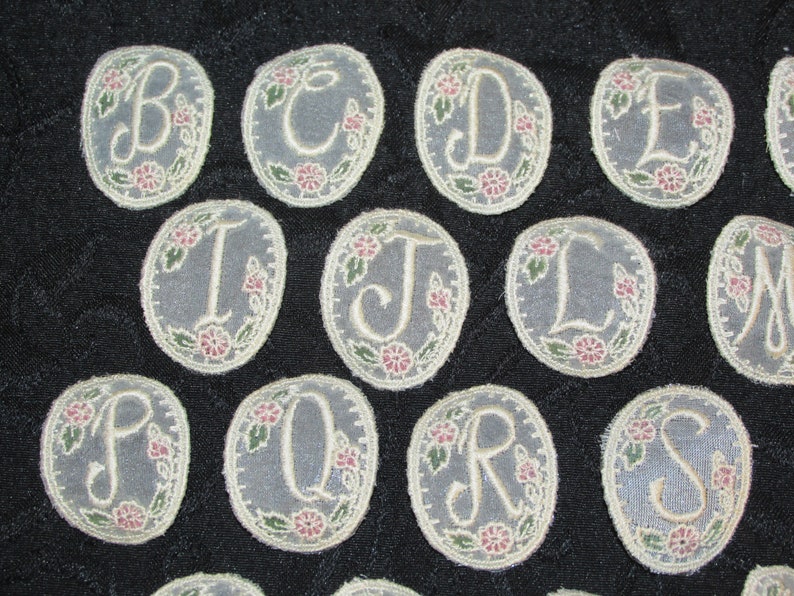 May include: A set of 13 oval-shaped fabric patches with embroidered letters of the alphabet, A through M, in a cream color. The letters are surrounded by a floral design with green leaves and pink flowers.