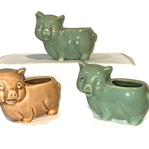 May include: Three ceramic pig planters, two in a light green glaze and one in a light brown glaze. Each planter has a small opening at the top for planting.
