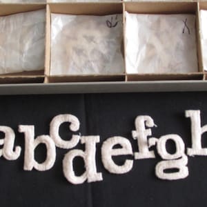 May include: White embroidered lowercase letters a through h are displayed on a black surface. The letters are in a serif font. Above the letters are several cardboard boxes with tissue paper inside.