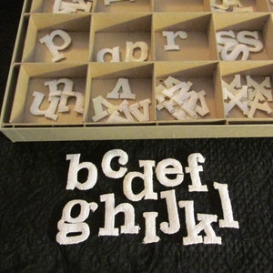 May include: A white plastic storage box with compartments filled with white felt letters. The letters are in various sizes and include the alphabet from a to z. The letters are arranged in alphabetical order. The box is sitting on a black surface.