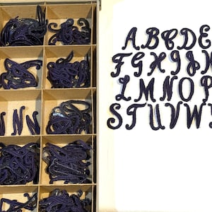 May include: A set of 26 black felt letters in a cursive font, arranged in alphabetical order. The letters are displayed on a white background. The letters are in a box with compartments filled with black felt scraps.