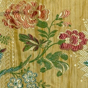 May include: A golden yellow fabric featuring an intricate embroidered floral pattern. The design includes large, multi-layered blossoms in shades of coral and deep pink, accompanied by green leaves, stems, and smaller light blue flowers.