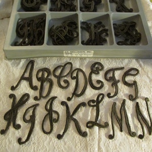 May include: A set of brown felt letters in a cursive font. The letters are A, B, C, D, E, F, G, H, I, J, K, L, M, and N. The letters are arranged in two rows. The letters are in a plastic container with the text "J.C. NELLISSEN, INC. 3008" on the label.