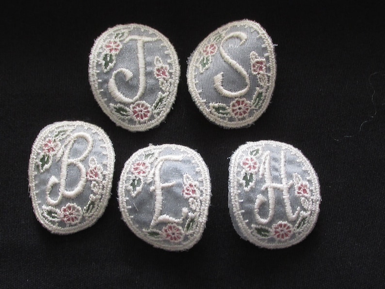 May include: Six embroidered fabric patches with white letters on a light blue background. Each patch has a white letter surrounded by a floral design with pink and green flowers.